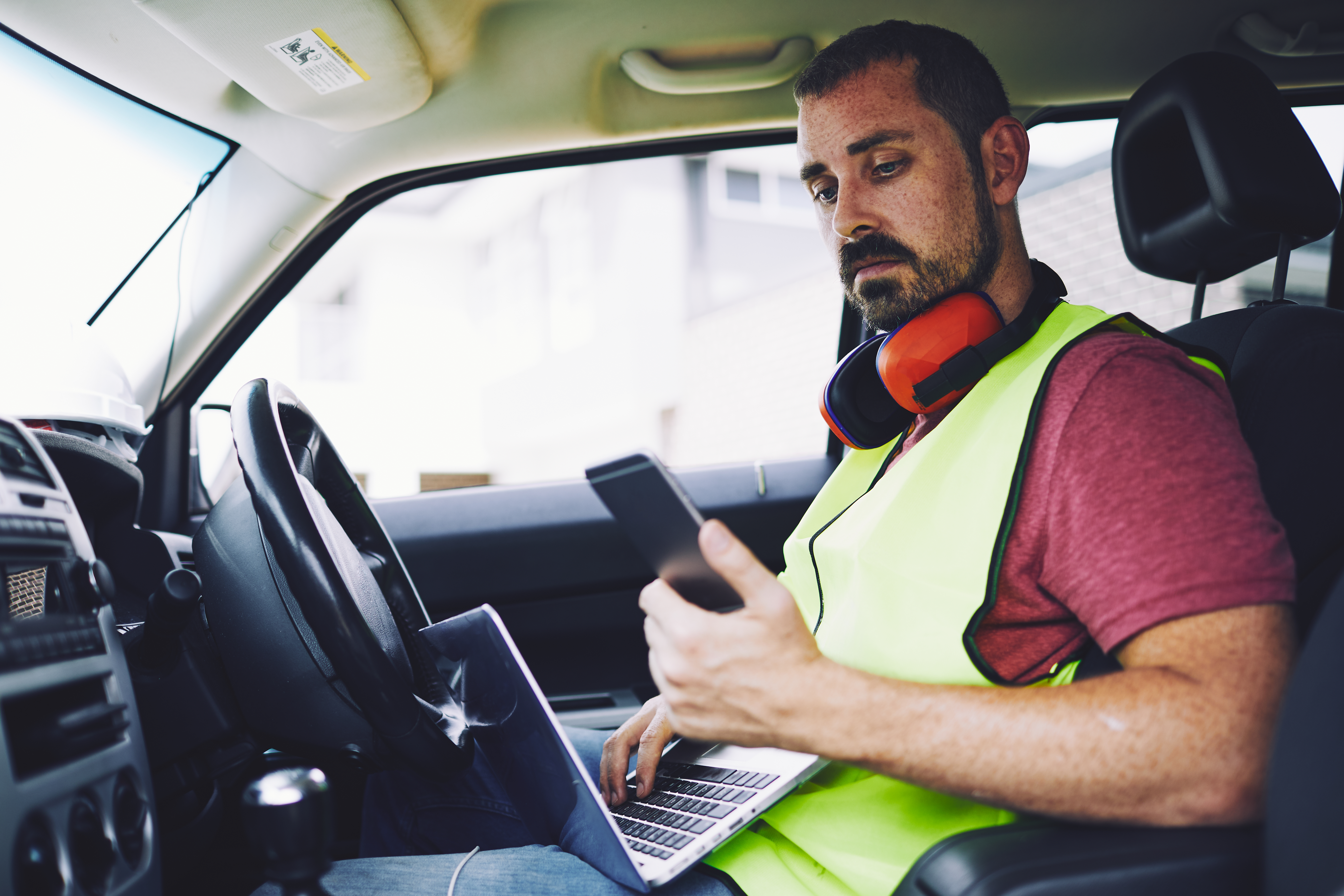 photo of contractor in vehicle looking at smartphone with laptop in his lap